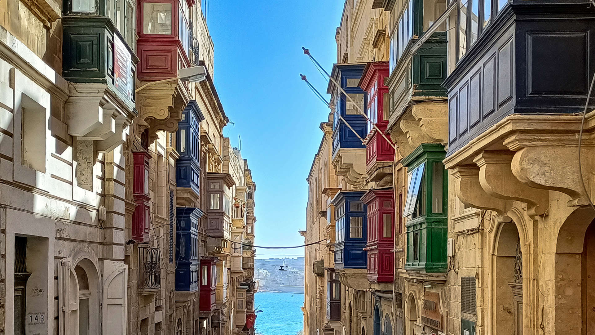 View in between buildings in Valetta (Malta) with a small piece of the ocean seen in the distance. Colourful balconies on the houses